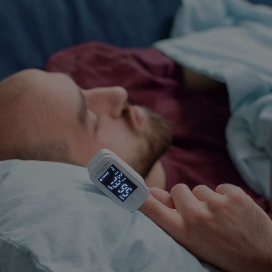 Person lying in bed with a pulse oximeter on their finger, showing heart rate and oxygen levels.